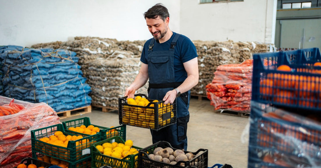 A worker carries a crate of lemons among stacked bins and pallets filled with assorted fresh produce.