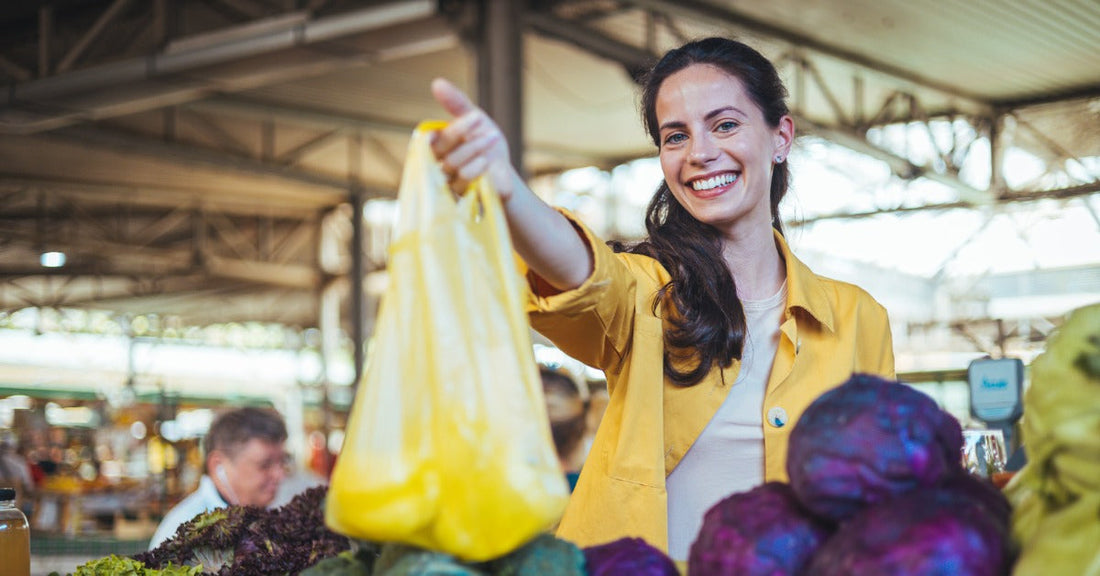 A smiling woman holds up a yellow produce bag behind a market stall piled with peppers, cabbage, and garlic.