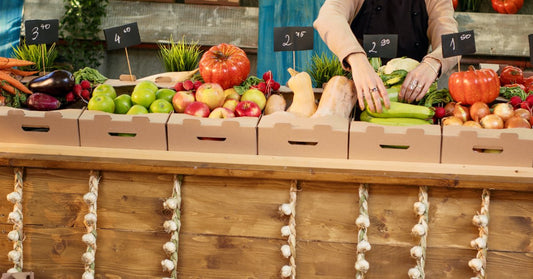 A vendor arranges fresh vegetables and fruit in cardboard boxes across a colorful farmers market stand.
