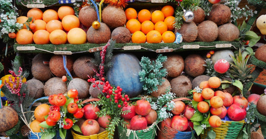 Colorful display of fresh oranges, apples, pomegranates, and coconuts arranged in decorative baskets with festive greenery and ornaments.