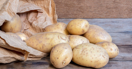 A brown paper bag is laying on a wooden table as fresh, light brown potatoes spill out of it onto the table.