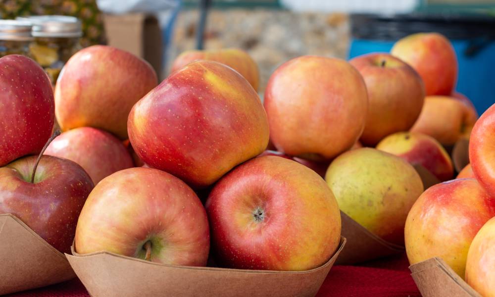 Large, red Fuji apples piled into paper cartons sit on a table for sale at a farmers market in front of other produce.