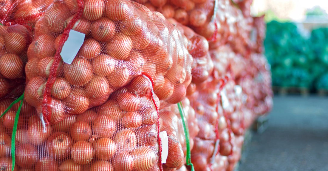 Stacked bags of yellow onions packed in red mesh produce bags sit on top of wooden pallets, awaiting pickup.