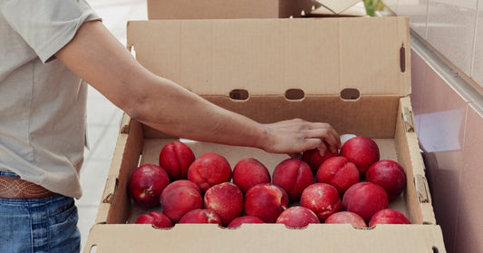 A person hand-selects nectarines to transfer from a large cardboard box sitting on a cart into a shallow box.