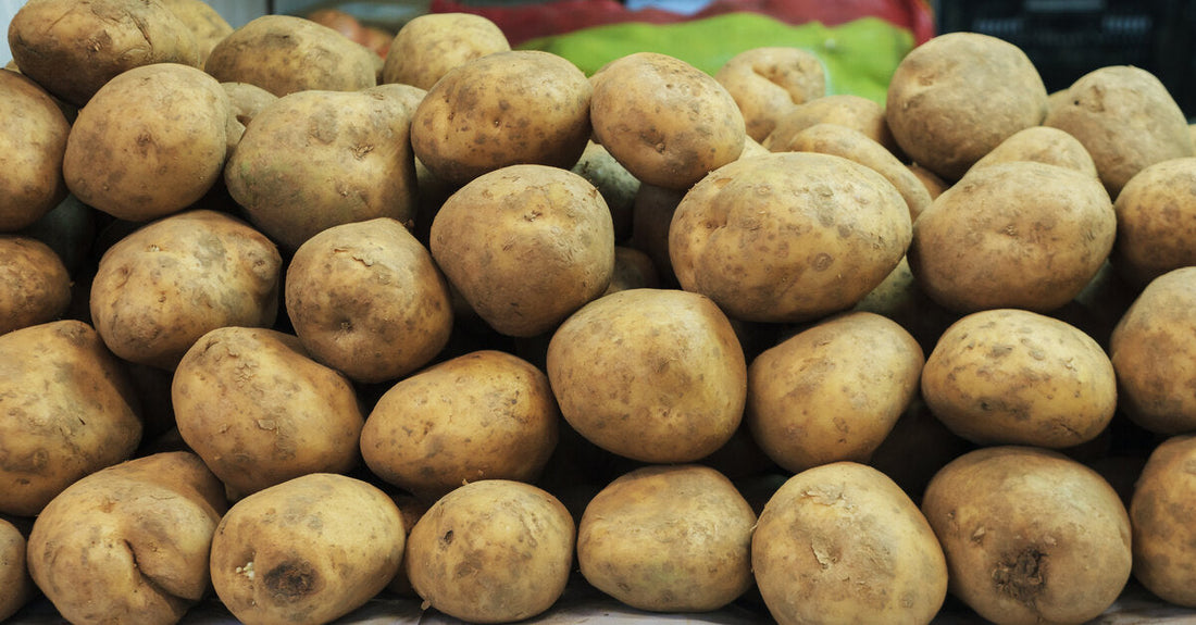 A large mound of potatoes sits on a counter covered in white paper. Crates of produce sit in the background.