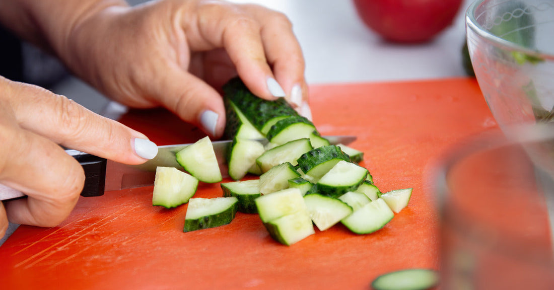 A close-up view of a woman's hands chopping a cucumber on a red plastic cutting board. A glass bowl and a tomato sit nearby.