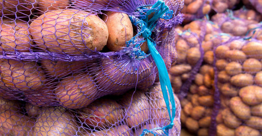 Large purple and blue mesh bags of fresh potatoes are stacked on top of one another in various piles.