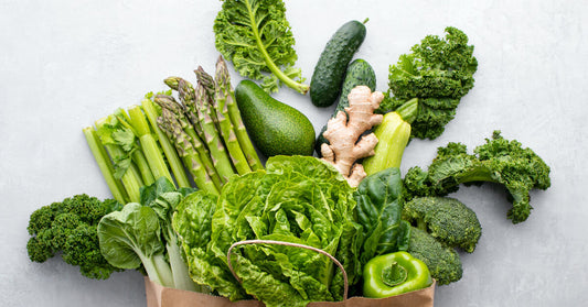 A brown paper grocery bag filled with an assortment of green, leafy vegetables lies on a gray background.