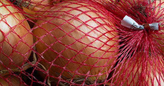 A red mesh bag holds eight large yellow onions. It's tied with a small white tag with black text at one end.