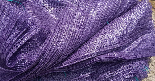 A large stack of purple mesh packaging nets is piled up on a concrete surface. The material is shiny and folded over on itself.