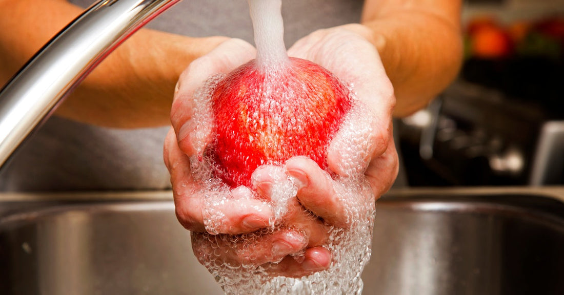 A pair of hands washes a vibrant red apple under running water from a sleek kitchen faucet, with water splashing below.