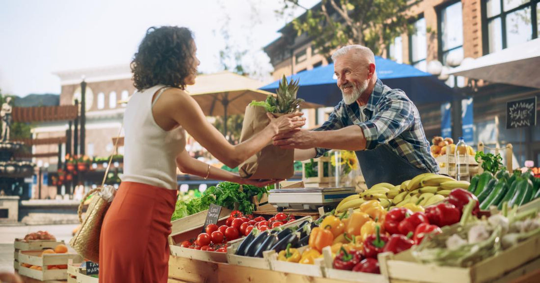 A vendor hands a brown bag of fresh produce to a customer at a vibrant farmers market filled with fruits and vegetables.