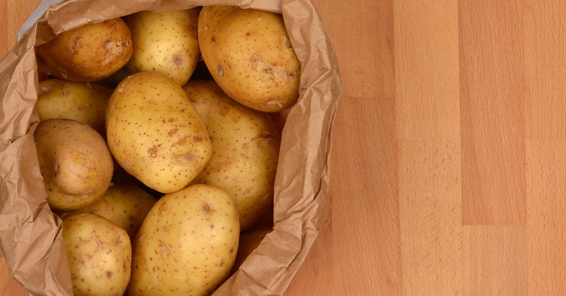 A top-down view of light yellow potatoes with small brown spots in a brown paper bag on a light wooden surface.