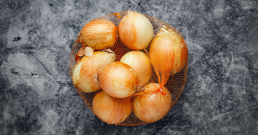 A mesh bag filled with yellow onions sits on a dark, textured surface. The onions vary in size with golden-brown skins.