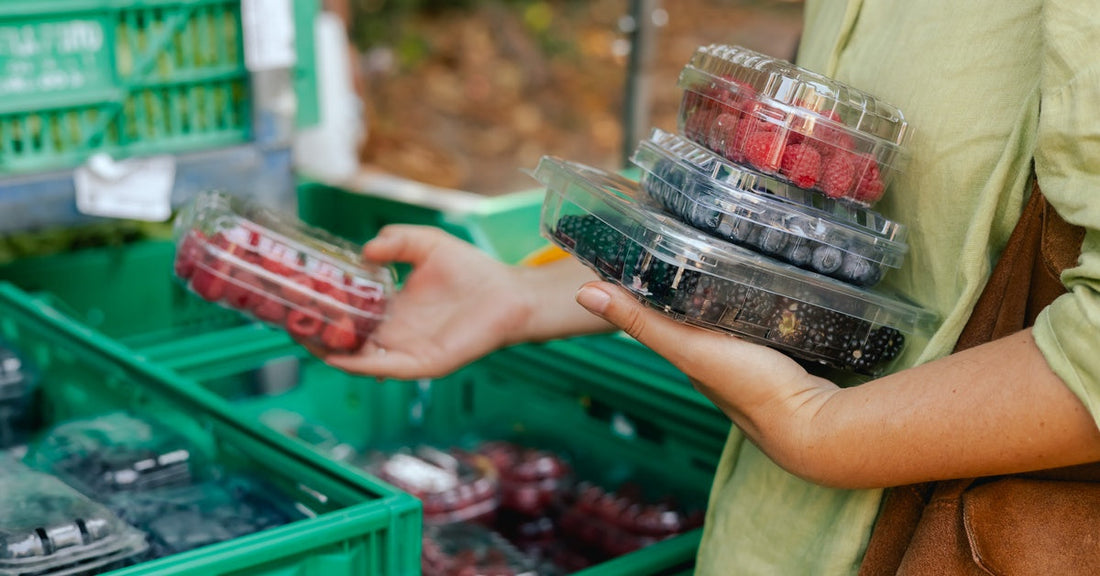 A person wearing a green shirt holds three containers of berries as they look through produce at a farmers market.