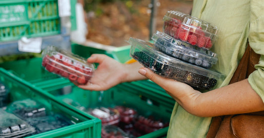 A person wearing a green shirt holds three containers of berries as they look through produce at a farmers market.