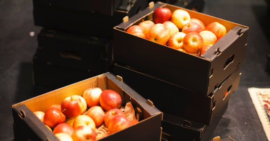 Stacked black cardboard boxes hold red and yellow apples on a dark warehouse floor under warm lighting.
