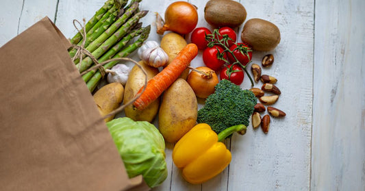 Fresh vegetables and fruits spill from a large brown paper grocery bag onto a white wooden table surface.