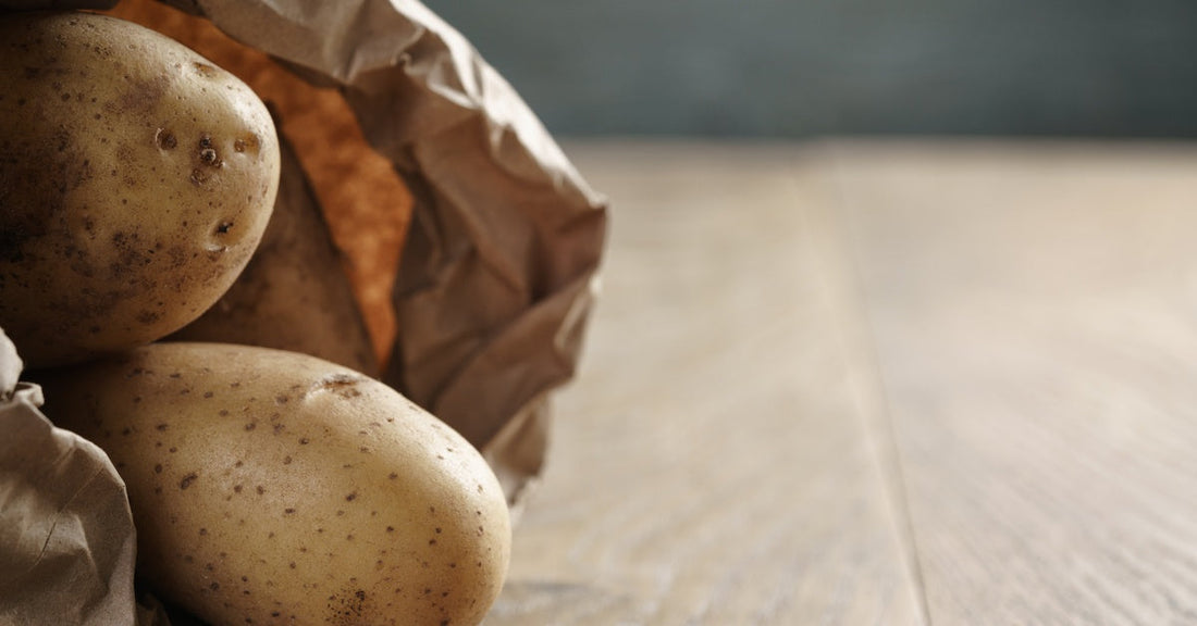 Potatoes spill out of a brown paper bag on a wooden table, with soft light highlighting their rough skins.