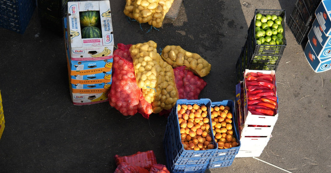 An overhead view shows crates and mesh bags filled with potatoes, peppers, apples, and citrus at a distribution center.