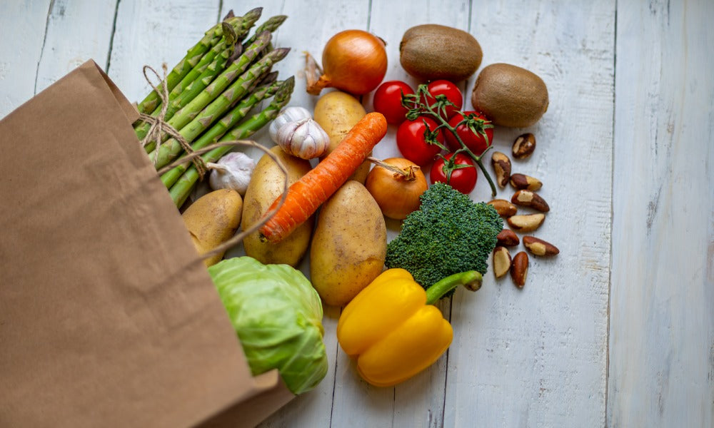 A brown paper bag spills asparagus, potatoes, cabbage, tomatoes, and other produce across a gray table.