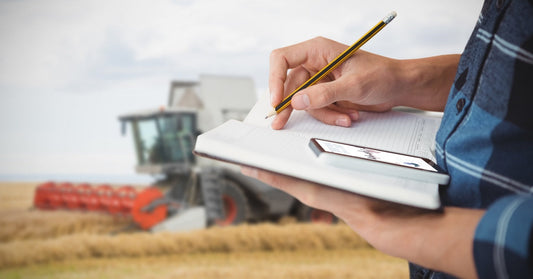 A farmer writes notes on a clipboard while a combine harvester works through a golden field in the background.
