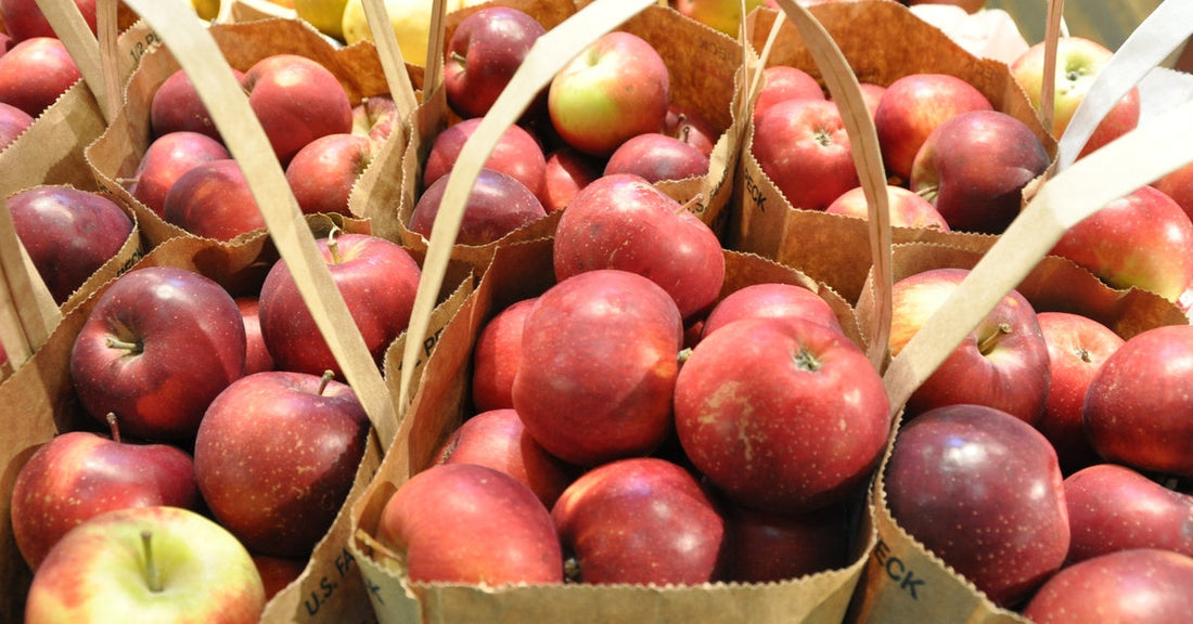 A close-up of several brown paper bags filled with red apples. Stacks of red and yellow apples sit behind them.